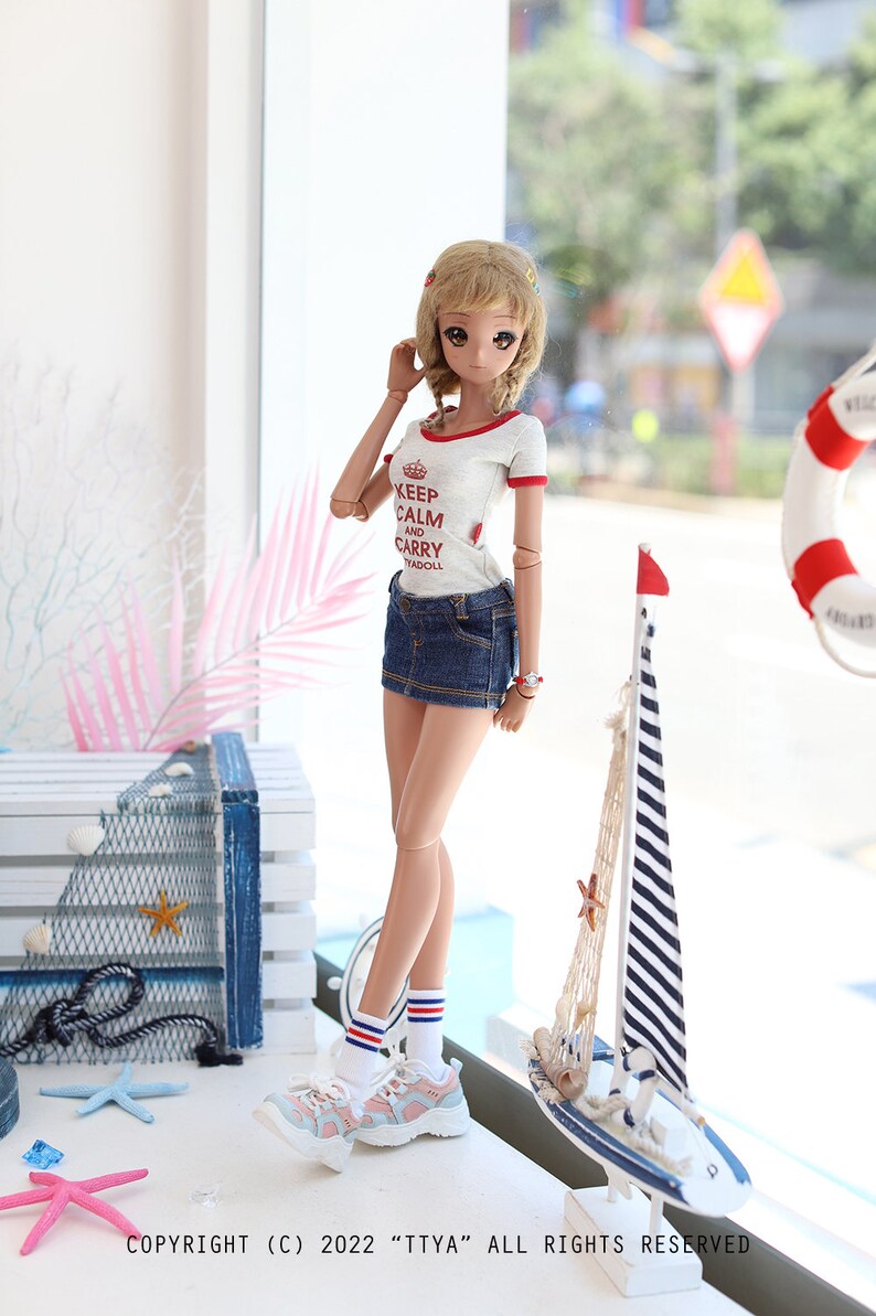 Puede incluir: Una mu&ntilde;eca con una camiseta blanca con texto rojo que dice "Keep Calm and Carry Yadoll", pantalones cortos de mezclilla azul, calcetines blancos con rayas rojas y azules y zapatillas de deporte rosa y blancas. La mu&ntilde;eca est&aacute; de pie frente a una ventana con un velero y un salvavidas visibles en el fondo.