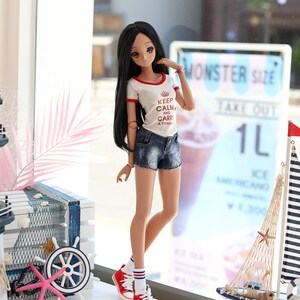 Puede incluir: Una mu&ntilde;eca con el pelo largo y negro que lleva una camiseta blanca con ribetes rojos y el texto "Keep Calm and Carry a Tiyado". Lleva pantalones cortos de mezclilla y calcetines a rayas rojas y blancas con zapatillas rojas. La mu&ntilde;eca est&aacute; de pie frente a una ventana con un cartel que dice "Monster Size Take Out 1L".