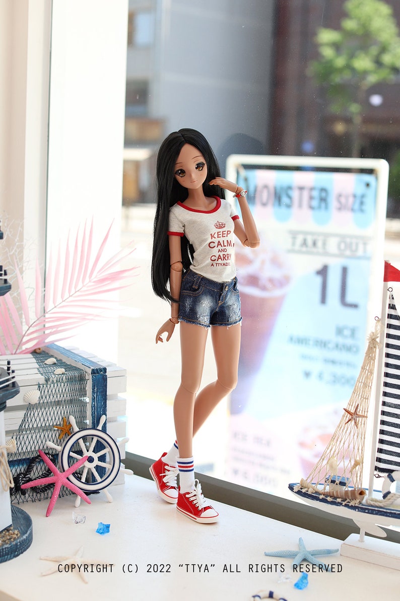 Puede incluir: Una mu&ntilde;eca con el pelo largo y negro que lleva una camiseta blanca con ribetes rojos y el texto "Keep Calm and Carry a Tiyado". Lleva pantalones cortos de mezclilla y calcetines a rayas rojas y blancas con zapatillas rojas. La mu&ntilde;eca est&aacute; de pie frente a una ventana con un cartel que dice "Monster Size Take Out 1L".