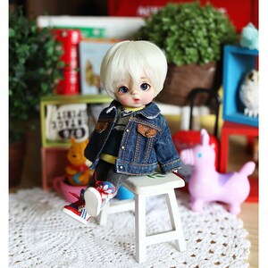 以下が含まれることがあります： 白い髪と青い目をした小さな人形が白いスツールに座っています。人形は青いデニムジャケット、黄色と緑のストライプのシャツ、青いジーンズを着ています。人形は白いレースのドイリーに座っています。