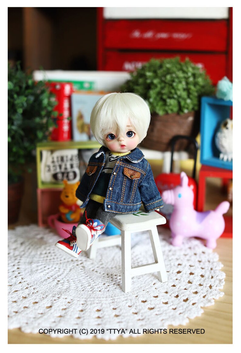 以下が含まれることがあります： 白い髪と青い目をした小さな人形が白いスツールに座っています。人形は青いデニムジャケット、黄色と緑のストライプのシャツ、青いジーンズを着ています。人形は白いレースのドイリーに座っています。