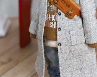 Puede incluir: Un abrigo gris claro con un suéter marrón debajo. El abrigo tiene botones grandes y una pequeña bolsa de cuero marrón con el texto "TTYA" se lleva en el pecho. El suéter tiene el texto "HIK SHI".