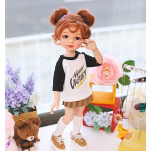 Puede incluir: Una mu&ntilde;eca con el pelo rojo en coletas que lleva una camiseta de b&eacute;isbol blanca y negra con las palabras "Honor Victory" impresas. La mu&ntilde;eca lleva una falda marr&oacute;n y calcetines blancos con zapatos marrones. La mu&ntilde;eca est&aacute; de pie sobre una superficie blanca con un oso de peluche y una maceta de flores en el fondo.