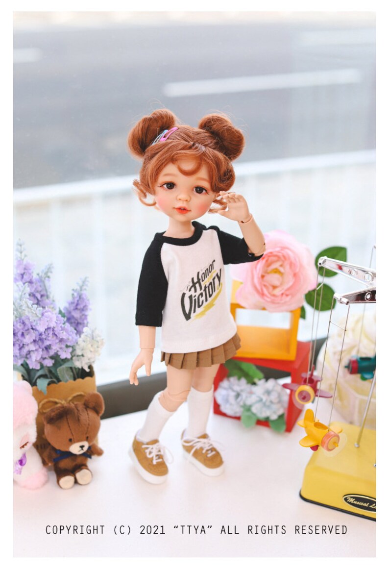 Puede incluir: Una mu&ntilde;eca con el pelo rojo en coletas que lleva una camiseta de b&eacute;isbol blanca y negra con las palabras "Honor Victory" impresas. La mu&ntilde;eca lleva una falda marr&oacute;n y calcetines blancos con zapatos marrones. La mu&ntilde;eca est&aacute; de pie sobre una superficie blanca con un oso de peluche y una maceta de flores en el fondo.