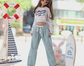Puede incluir: Una muñeca con una sudadera corta gris con "Collegiate" y "37" estampados, combinada con pantalones azul claro y zapatillas deportivas. Un velero en miniatura, un salvavidas y una tabla de surf con "Keep it Simple Surf More" están en el fondo.