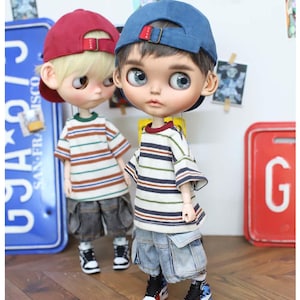 Può includere: Due bambole con magliette a righe, pantaloncini di jeans e berretti da baseball. Una bambola indossa un berretto rosso, l'altra un berretto blu. Entrambe le bambole indossano scarpe da ginnastica. Lo sfondo include una targa blu e un cartello rosso con la lettera "G".