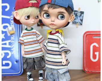 Puede incluir: Dos muñecos con camisetas a rayas, pantalones cortos de mezclilla y gorras de béisbol. Un muñeco lleva una gorra roja, el otro una gorra azul. Ambos muñecos llevan zapatillas deportivas. El fondo incluye una placa de matrícula azul y un letrero rojo con la letra "G".