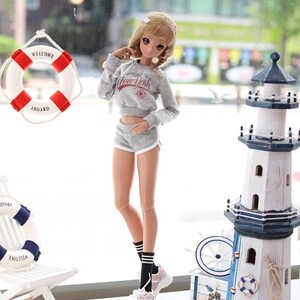May include: A doll wearing a gray sweatshirt with "New York" printed on it, gray shorts, black socks with white stripes, and light blue and pink sneakers. The doll is standing in front of a window with a life preserver and a miniature lighthouse.