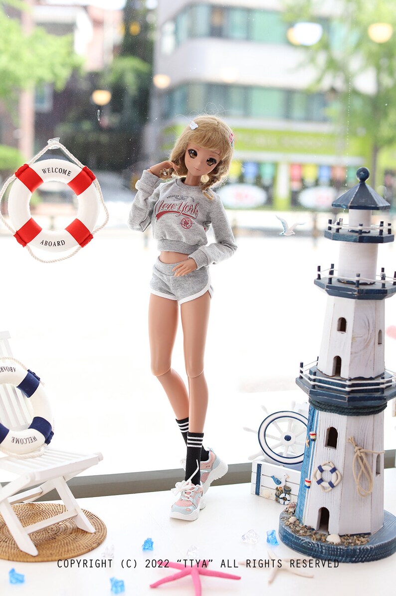 May include: A doll wearing a gray sweatshirt with "New York" printed on it, gray shorts, black socks with white stripes, and light blue and pink sneakers. The doll is standing in front of a window with a life preserver and a miniature lighthouse.