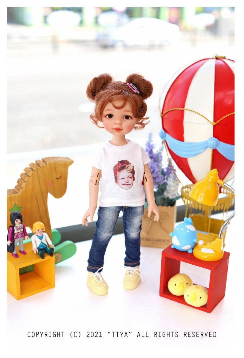 Puede incluir: Una mu&ntilde;eca con el pelo casta&ntilde;o en coletas que lleva una camiseta blanca con una imagen de una persona, pantalones vaqueros azules y zapatillas amarillas. La mu&ntilde;eca est&aacute; de pie delante de un caballo de balanc&iacute;n de madera y un globo aerost&aacute;tico rojo y blanco.