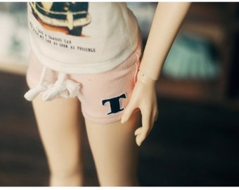Könnte beinhalten: Eine Puppe trägt ein weißes T-Shirt mit einem Autodesign und Text, kombiniert mit rosa Shorts. Die Shorts haben einen schwarzen "T"-Aufnäher. Das Outfit ist lässig und scheint für eine Puppe zu sein.