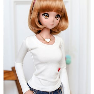 May include: A doll with short, brown hair wearing a white long-sleeved shirt, a denim skirt, and a red bandana headband. The doll is wearing a gold necklace with a white heart pendant.