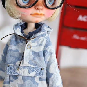 May include: A doll with blue eyes and blonde hair wearing a blue and white camouflage patterned jacket with a hood. The jacket has a pocket and a brown drawstring. The doll is wearing black glasses.
