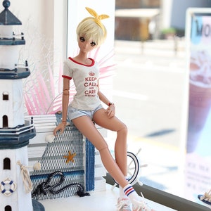 Puede incluir: Una mu&ntilde;eca con cabello rubio que lleva una camiseta gris con el texto "Keep Calm and Carry a Tiyada" en rojo. La mu&ntilde;eca lleva una diadema amarilla, pantalones cortos de mezclilla azul, calcetines blancos con rayas rojas y azules y zapatillas de deporte azul claro. La mu&ntilde;eca est&aacute; sentada en una repisa de madera blanca con una red azul y blanca detr&aacute;s. Hay un faro blanco y una estrella de mar rosa en la repisa.