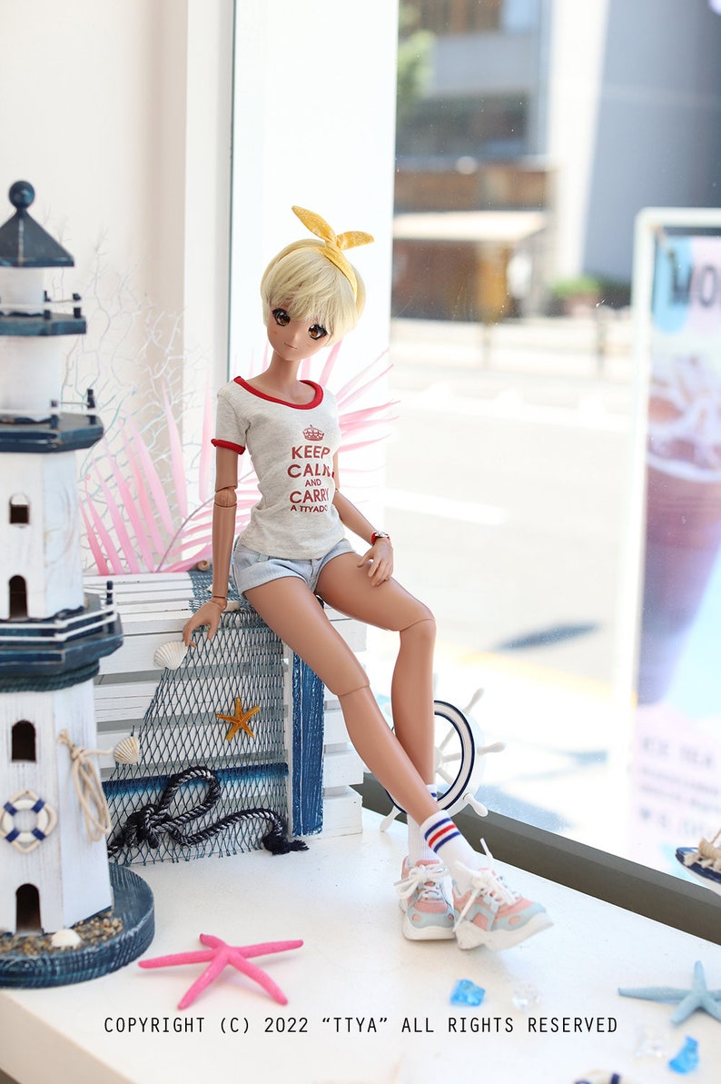 Puede incluir: Una mu&ntilde;eca con cabello rubio que lleva una camiseta gris con el texto "Keep Calm and Carry a Tiyada" en rojo. La mu&ntilde;eca lleva una diadema amarilla, pantalones cortos de mezclilla azul, calcetines blancos con rayas rojas y azules y zapatillas de deporte azul claro. La mu&ntilde;eca est&aacute; sentada en una repisa de madera blanca con una red azul y blanca detr&aacute;s. Hay un faro blanco y una estrella de mar rosa en la repisa.