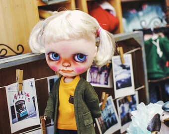 May include: A doll with blonde hair in pigtails, blue eyes, and rosy cheeks, wearing a mustard yellow t-shirt with a brown pocket, an olive green cardigan, dark blue cuffed jeans, and brown lace-up boots. The text COPYRIGHT(C)2016 “TTYA” ALL RIGHTS RESERVED is visible.