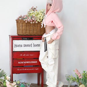 Puede incluir: Una sudadera rosa con capucha recortada con un cord&oacute;n blanco, puesta por una mu&ntilde;eca con cabello blanco y pantalones blancos. La mu&ntilde;eca est&aacute; de pie frente a un armario de madera rojo con las palabras "Home sweet home" escritas en &eacute;l.