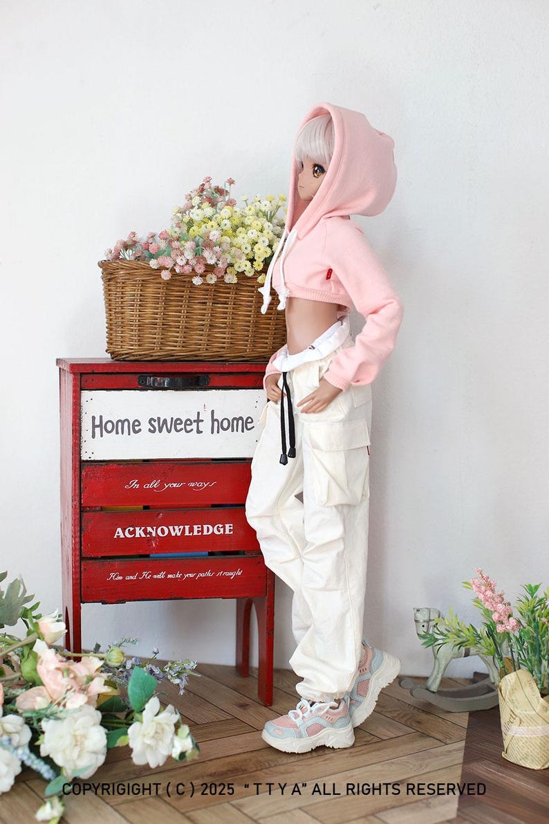 Puede incluir: Una sudadera rosa con capucha recortada con un cord&oacute;n blanco, puesta por una mu&ntilde;eca con cabello blanco y pantalones blancos. La mu&ntilde;eca est&aacute; de pie frente a un armario de madera rojo con las palabras "Home sweet home" escritas en &eacute;l.