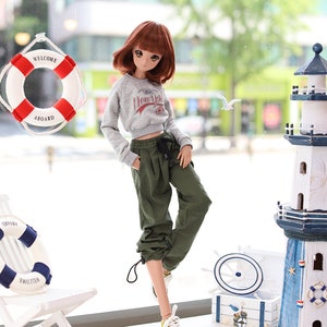 May include: A doll wearing a grey sweatshirt with the text "New York" on it, green cargo pants, and white sneakers. The doll is standing in front of a window with a life preserver and a lighthouse in the background.