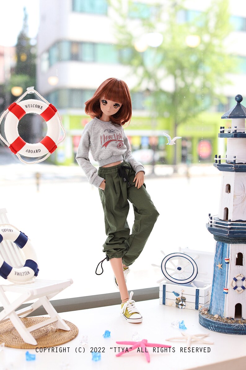 May include: A doll wearing a grey sweatshirt with the text "New York" on it, green cargo pants, and white sneakers. The doll is standing in front of a window with a life preserver and a lighthouse in the background.
