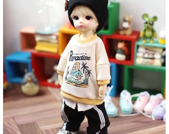 May include: A ball-jointed doll with blonde hair wearing a black beanie, a cream-colored long-sleeve sweatshirt with yellow ribbed trim and a graphic of a bird with the text "Living Life In Paradise," and black track pants with white side stripes.