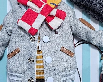 Puede incluir: Una chaqueta bomber gris en miniatura con parches marrones y botones blancos. Una bufanda a cuadros rojos y blancos y una bufanda amarilla y azul alrededor del cuello. Se ve una camisa a rayas amarillas y blancas.