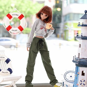 May include: A doll wearing a grey sweatshirt with a graphic print and green cargo pants. The doll is holding a yellow mug and is standing in front of a window with a life preserver hanging on it. The doll is wearing yellow sneakers.