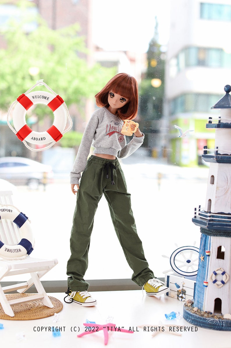 May include: A doll wearing a grey sweatshirt with a graphic print and green cargo pants. The doll is holding a yellow mug and is standing in front of a window with a life preserver hanging on it. The doll is wearing yellow sneakers.