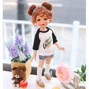 Puede incluir: Una mu&ntilde;eca con el pelo casta&ntilde;o en coletas que lleva una camiseta de b&eacute;isbol blanca y negra con el texto "Honor Victory". La mu&ntilde;eca lleva pantalones cortos de mezclilla y calcetines blancos con zapatos marrones. La mu&ntilde;eca est&aacute; de pie sobre una superficie blanca con un oso de peluche, una flor rosa y un caballo de balanc&iacute;n de madera.