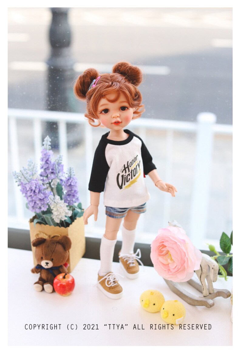 Puede incluir: Una mu&ntilde;eca con el pelo casta&ntilde;o en coletas que lleva una camiseta de b&eacute;isbol blanca y negra con el texto "Honor Victory". La mu&ntilde;eca lleva pantalones cortos de mezclilla y calcetines blancos con zapatos marrones. La mu&ntilde;eca est&aacute; de pie sobre una superficie blanca con un oso de peluche, una flor rosa y un caballo de balanc&iacute;n de madera.