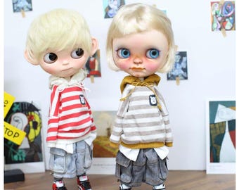 May include: Two Blythe dolls with blonde hair are dressed in casual outfits. One doll wears a red and white striped hoodie, denim shorts, and red and black sneakers. The other doll wears a gray and white striped hoodie, denim shorts, and black and white sneakers.