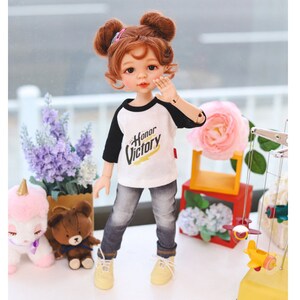 Puede incluir: Una mu&ntilde;eca con el pelo rojo en coletas que lleva una camiseta de b&eacute;isbol blanca y negra con el texto "Honor Victory" en ella. La mu&ntilde;eca lleva pantalones vaqueros azules y zapatillas amarillas. La mu&ntilde;eca est&aacute; de pie sobre una superficie blanca con un oso de peluche marr&oacute;n y un juguete de unicornio rosa en el fondo.