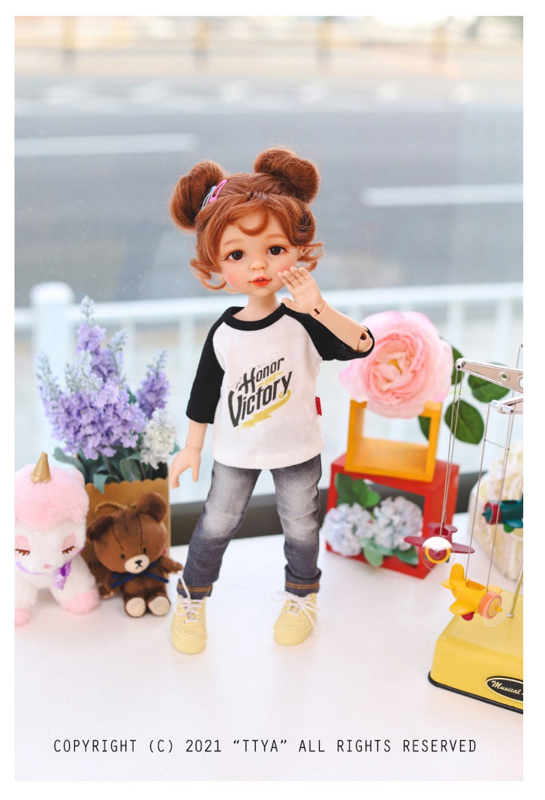 Puede incluir: Una mu&ntilde;eca con el pelo rojo en coletas que lleva una camiseta de b&eacute;isbol blanca y negra con el texto "Honor Victory" en ella. La mu&ntilde;eca lleva pantalones vaqueros azules y zapatillas amarillas. La mu&ntilde;eca est&aacute; de pie sobre una superficie blanca con un oso de peluche marr&oacute;n y un juguete de unicornio rosa en el fondo.