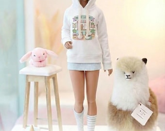 Puede incluir: Una sudadera con capucha blanca con estampado personalizado, combinada con una falda azul claro y calcetines a rayas, diseñada para una muñeca SD13. La sudadera con capucha presenta un gráfico colorido con temática de café. Un conejito de peluche y una alpaca también están en la imagen.