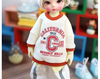 May include: A ball-jointed doll wearing a white beanie with a red accent, a cream-colored sweatshirt with orange trim and red text reading CALIFORNIA LOS ANGELES CITY BEVERLY HILLS ATHLETIC DEPARTMENT, and white sweatpants with black side stripes.