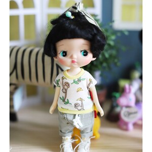May include: A small doll with black hair and blue eyes wearing a white t-shirt with a monkey and banana print, grey pants, and white sneakers. The doll is standing on a wooden surface.