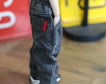 Puede incluir: Pantalones vaqueros grises oscuros con una etiqueta roja "TTYA". Los pantalones tienen cintura elástica y bolsillos. Los pantalones para muñecas se usan con zapatillas deportivas blancas y negras.
