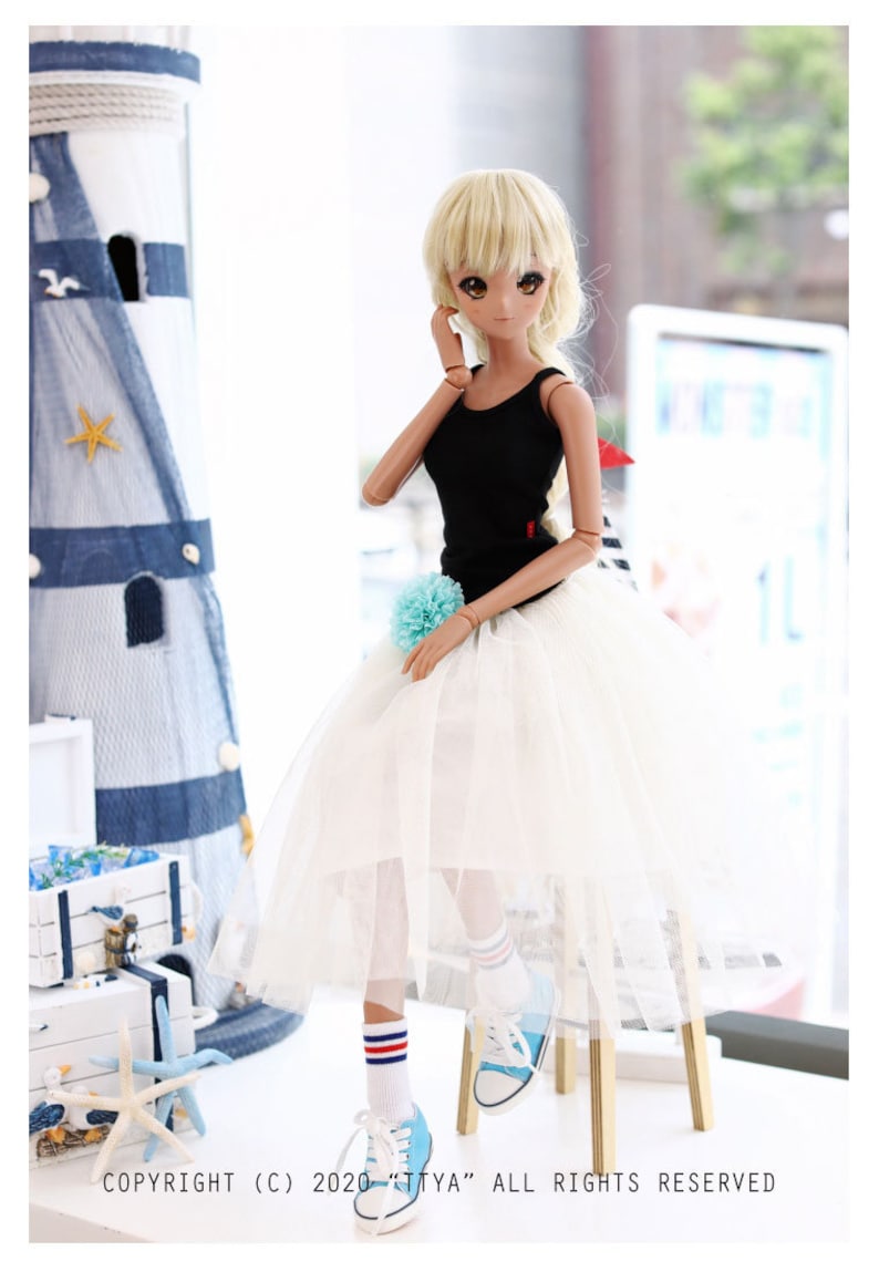 K&ouml;nnte beinhalten: Eine Puppe tr&auml;gt ein schwarzes Tanktop, einen wei&szlig;en T&uuml;llrock, blau-wei&szlig; gestreifte Socken und blaue Turnschuhe. Die Puppe hat blondes Haar und sitzt auf einem Holzhocker. Die Puppe steht vor einem Fenster mit einem blau-wei&szlig; gestreiften Vorhang.