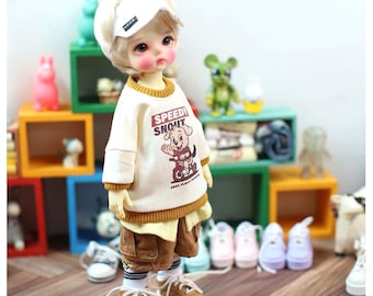 May include: A ball-jointed doll with blonde hair wearing a white visor, a cream-colored long-sleeve shirt with a brown graphic of a dog on a bicycle and the text "SPEEDY SNOUT", brown cargo shorts, white socks with black stripes, and brown sneakers.
