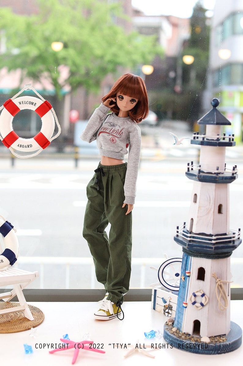 May include: A doll wearing a grey cropped sweatshirt with the text "New York" on the front, green cargo pants, and white sneakers. The doll is standing in front of a window with a life preserver and a miniature lighthouse.