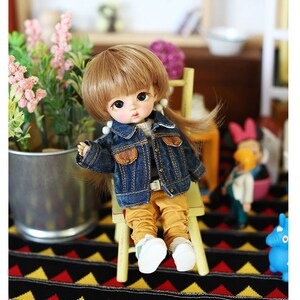 以下が含まれることがあります： 茶色の髪の毛をした小さな人形が、青いデニムジャケット、黄色のズボン、白い靴を履いて、黄色の椅子に座っています。人形は黒と黄色の模様の敷物の上に座っています。