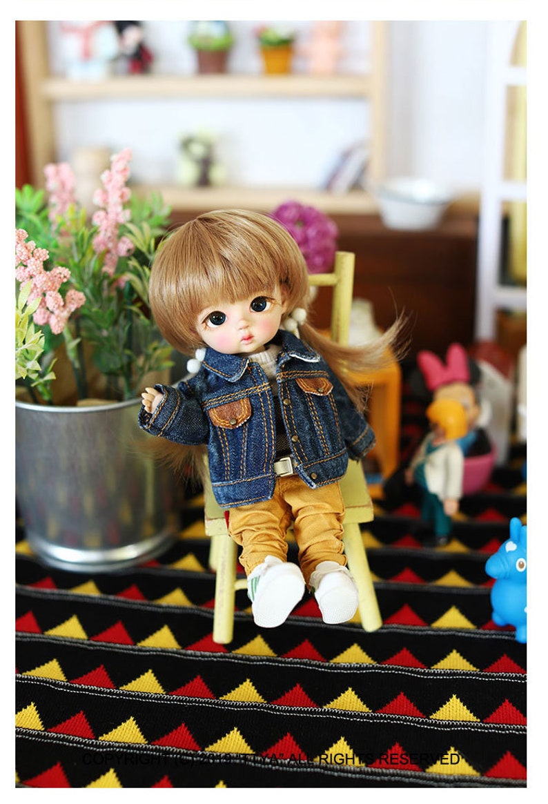 以下が含まれることがあります： 茶色の髪の毛をした小さな人形が、青いデニムジャケット、黄色のズボン、白い靴を履いて、黄色の椅子に座っています。人形は黒と黄色の模様の敷物の上に座っています。
