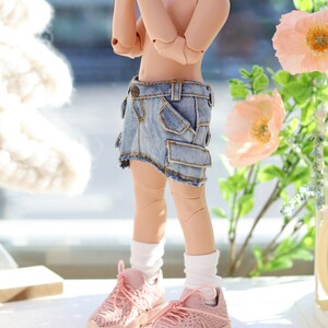 Pode incluir: Uma boneca vestindo uma saia de ganga com um bolso cargo, meias brancas e t&ecirc;nis rosa. A boneca tem cabelo rosa e est&aacute; em p&eacute; sobre uma superf&iacute;cie branca.