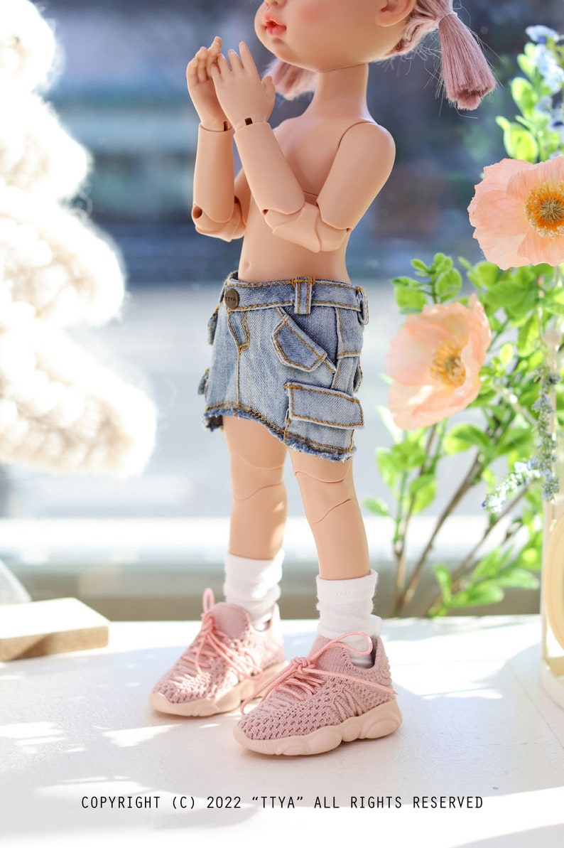 Pode incluir: Uma boneca vestindo uma saia de ganga com um bolso cargo, meias brancas e t&ecirc;nis rosa. A boneca tem cabelo rosa e est&aacute; em p&eacute; sobre uma superf&iacute;cie branca.