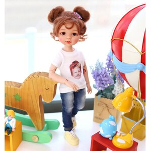 Puede incluir: Una mu&ntilde;eca con el pelo casta&ntilde;o en coletas que lleva una camiseta blanca con una foto de una mujer, pantalones vaqueros azules y zapatos amarillos. La mu&ntilde;eca est&aacute; de pie junto a un caballo de balanc&iacute;n de madera y una caja de madera roja con una l&aacute;mpara amarilla y una figurita de nube azul en la parte superior.