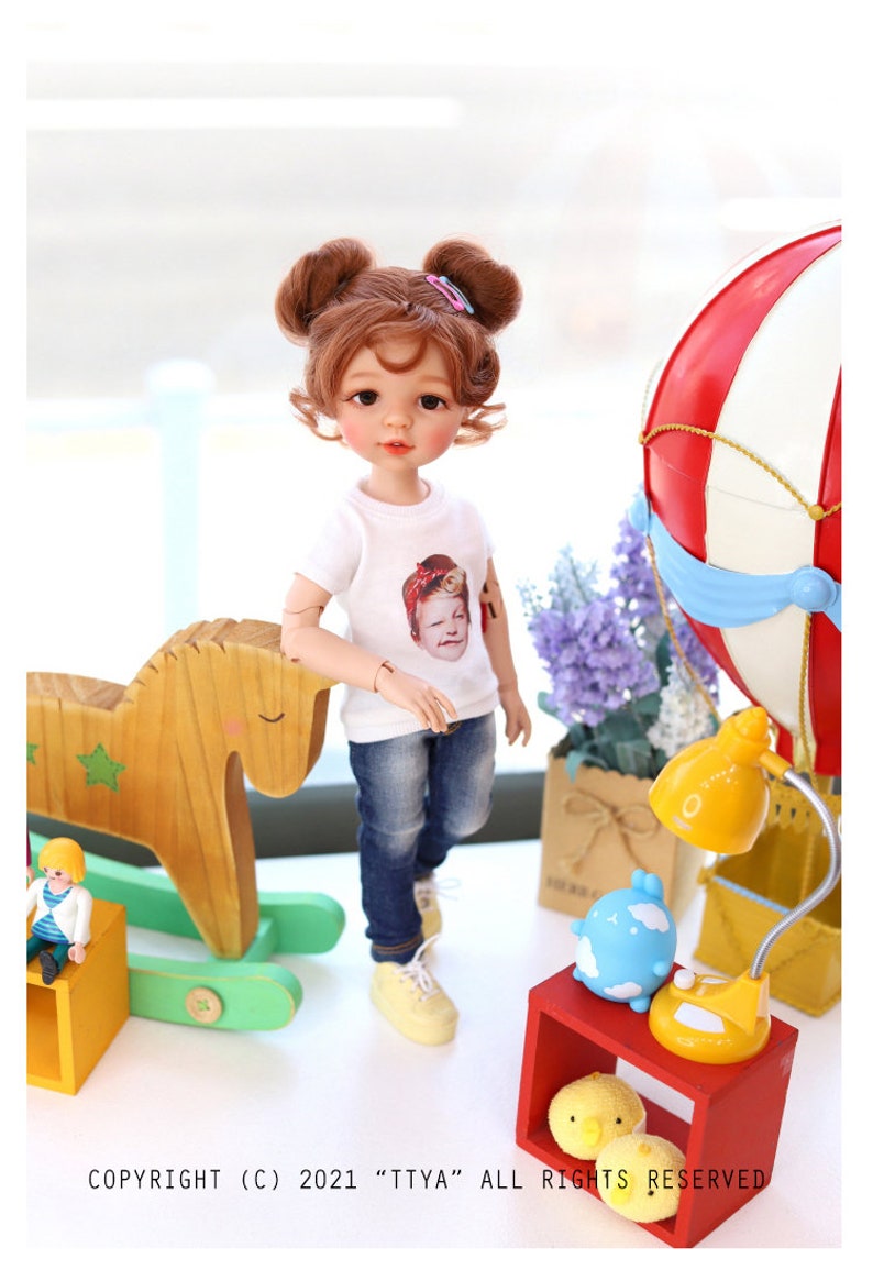 Puede incluir: Una mu&ntilde;eca con el pelo casta&ntilde;o en coletas que lleva una camiseta blanca con una foto de una mujer, pantalones vaqueros azules y zapatos amarillos. La mu&ntilde;eca est&aacute; de pie junto a un caballo de balanc&iacute;n de madera y una caja de madera roja con una l&aacute;mpara amarilla y una figurita de nube azul en la parte superior.