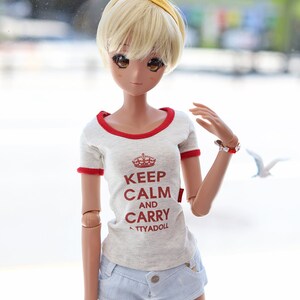 Puede incluir: Una mu&ntilde;eca con cabello rubio que lleva una camiseta blanca con ribetes rojos y el texto "Keep Calm and Carry ATTYADOLL" en rojo. La mu&ntilde;eca tambi&eacute;n lleva pantalones cortos de mezclilla azul y una diadema amarilla.