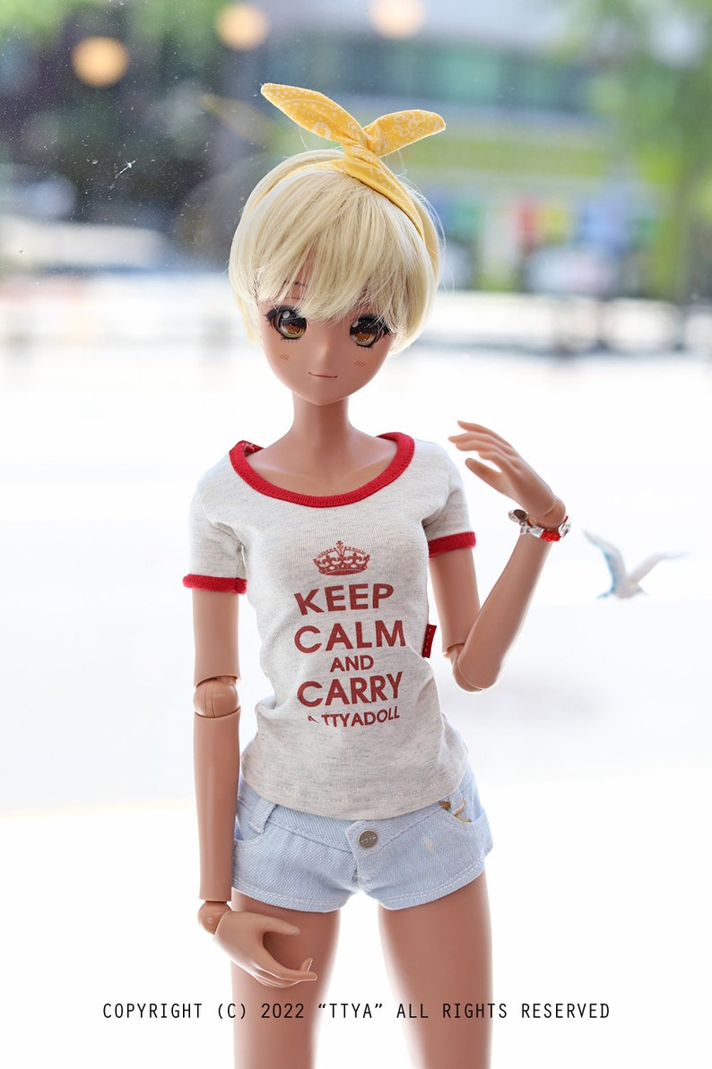 Puede incluir: Una mu&ntilde;eca con cabello rubio que lleva una camiseta blanca con ribetes rojos y el texto "Keep Calm and Carry ATTYADOLL" en rojo. La mu&ntilde;eca tambi&eacute;n lleva pantalones cortos de mezclilla azul y una diadema amarilla.