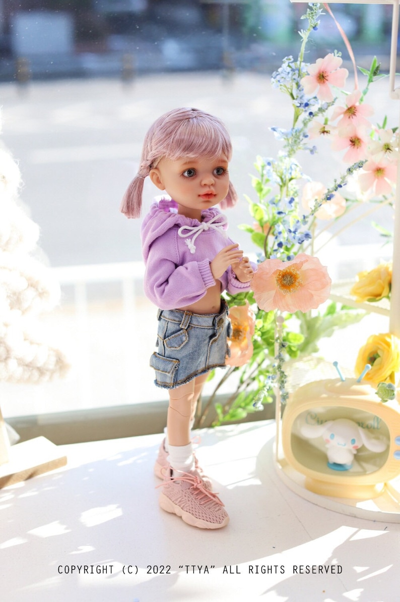 Pode incluir: Uma boneca rosa com cabelo rosa em tran&ccedil;as usando um moletom roxo com capuz e uma saia jeans azul. A boneca est&aacute; segurando uma flor rosa e est&aacute; em p&eacute; em uma superf&iacute;cie branca. A boneca est&aacute; usando t&ecirc;nis rosa. COPYRIGHT (C) 2022 "TTYA" ALL RIGHTS RESERVED