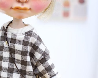 Puede incluir: Una muñeca con grandes ojos azules, cabello rubio y mejillas rosadas. La muñeca lleva un suéter a cuadros marrón y blanco con un pequeño bolso de mano beige. El atuendo de la muñeca tiene una estética vintage.